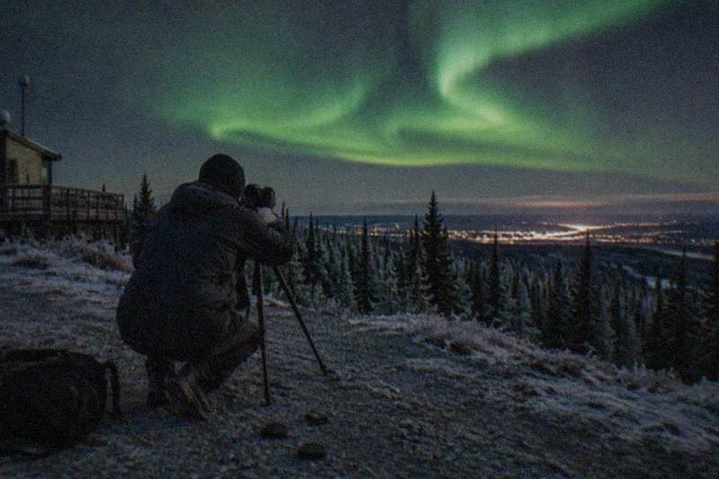 A photographer with a tripod aimed at the northern lights over the hills near Fairbanks, Alaska
